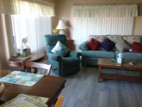 Ein Wohnzimmer mit Sofa, Sessel und Couchtisch. Auf einem Beistelltisch steht eine Lampe und eine Vase mit Blumen.