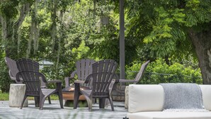 Een terras met Adirondack-stoelen en een witte bank, omgeven door weelderig groen en bomen.