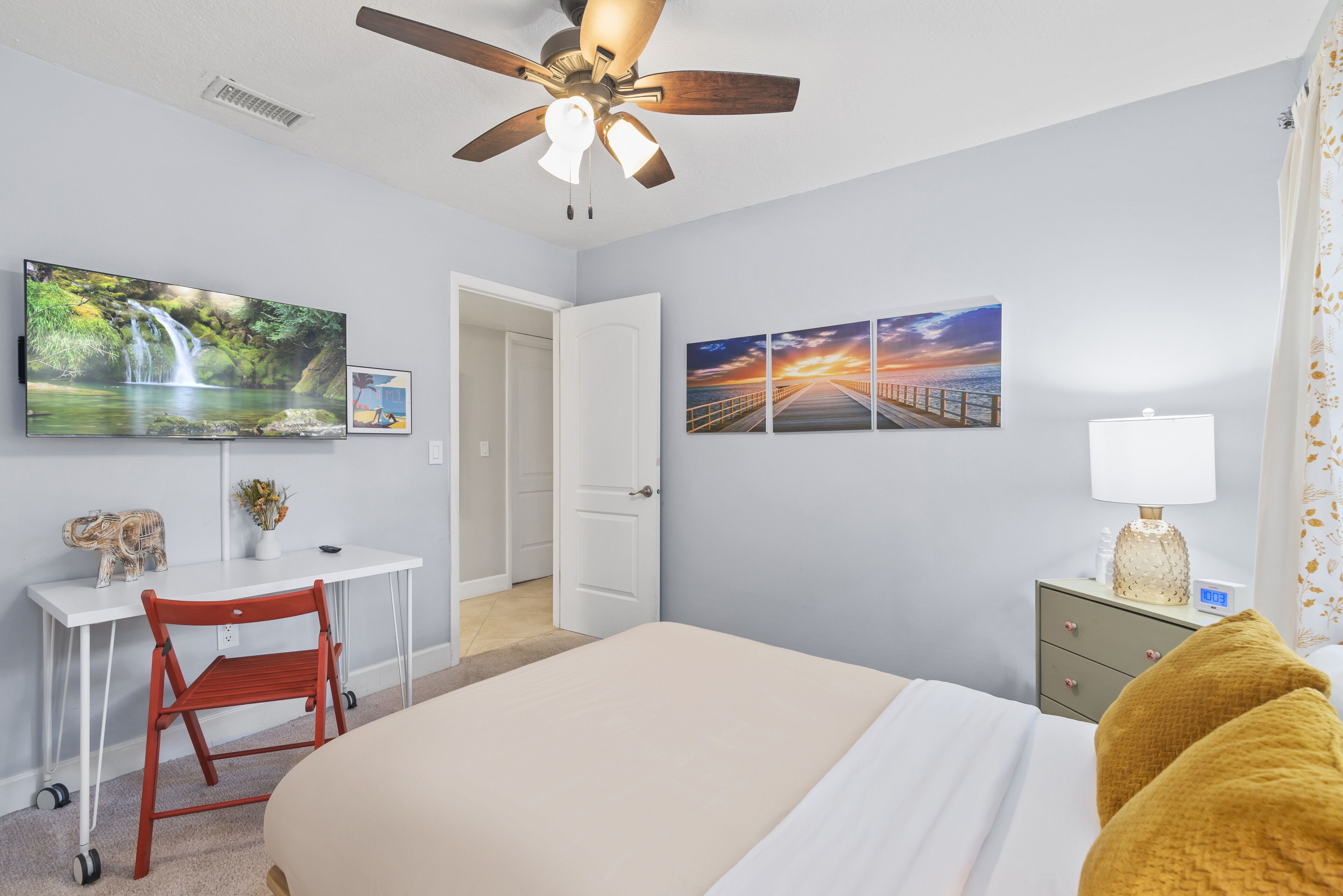 A bedroom with a bed, a desk, a chair, a TV, and artwork on the wall.