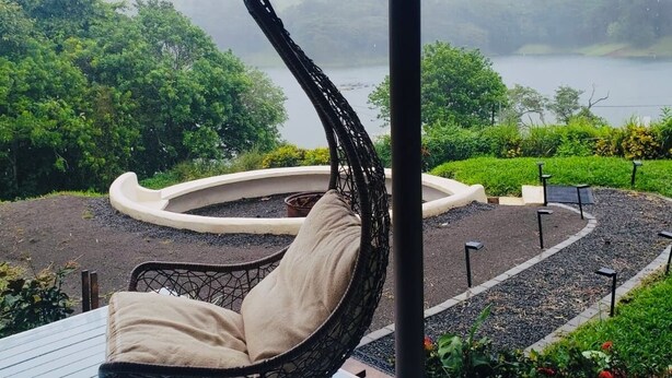 A hanging chair on a wooden deck overlooking a lake with a garden and a circular seating area.