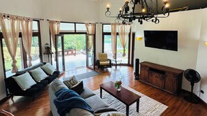 A spacious living room with a large sofa, a coffee table, and a TV mounted on the wall. There are curtains on the windows, and a chandelier hangs from the ceiling.