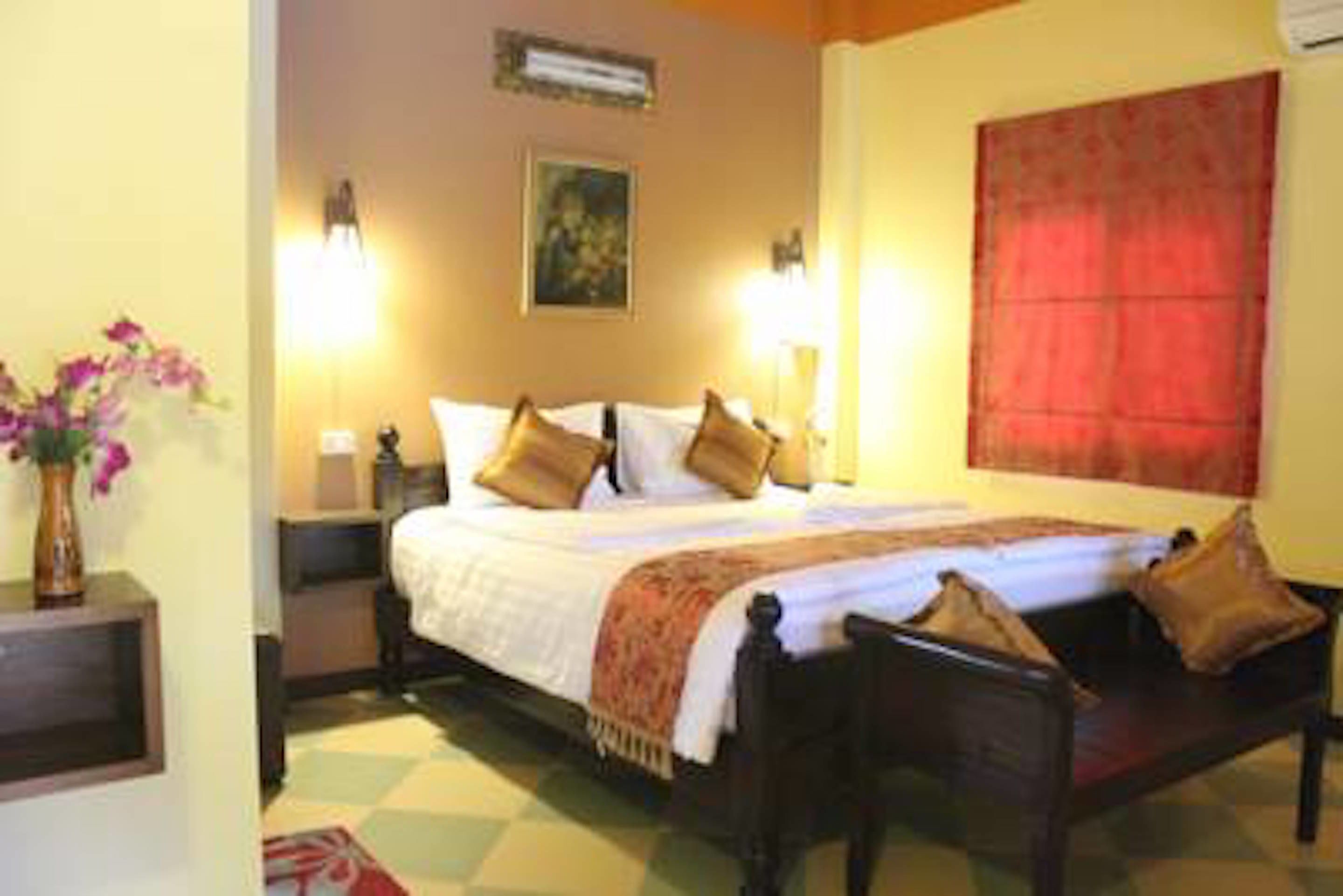 Habitación de hotel con cama, lámparas de noche, mesita de noche, un florero con flores y un ventanal con cortina roja.