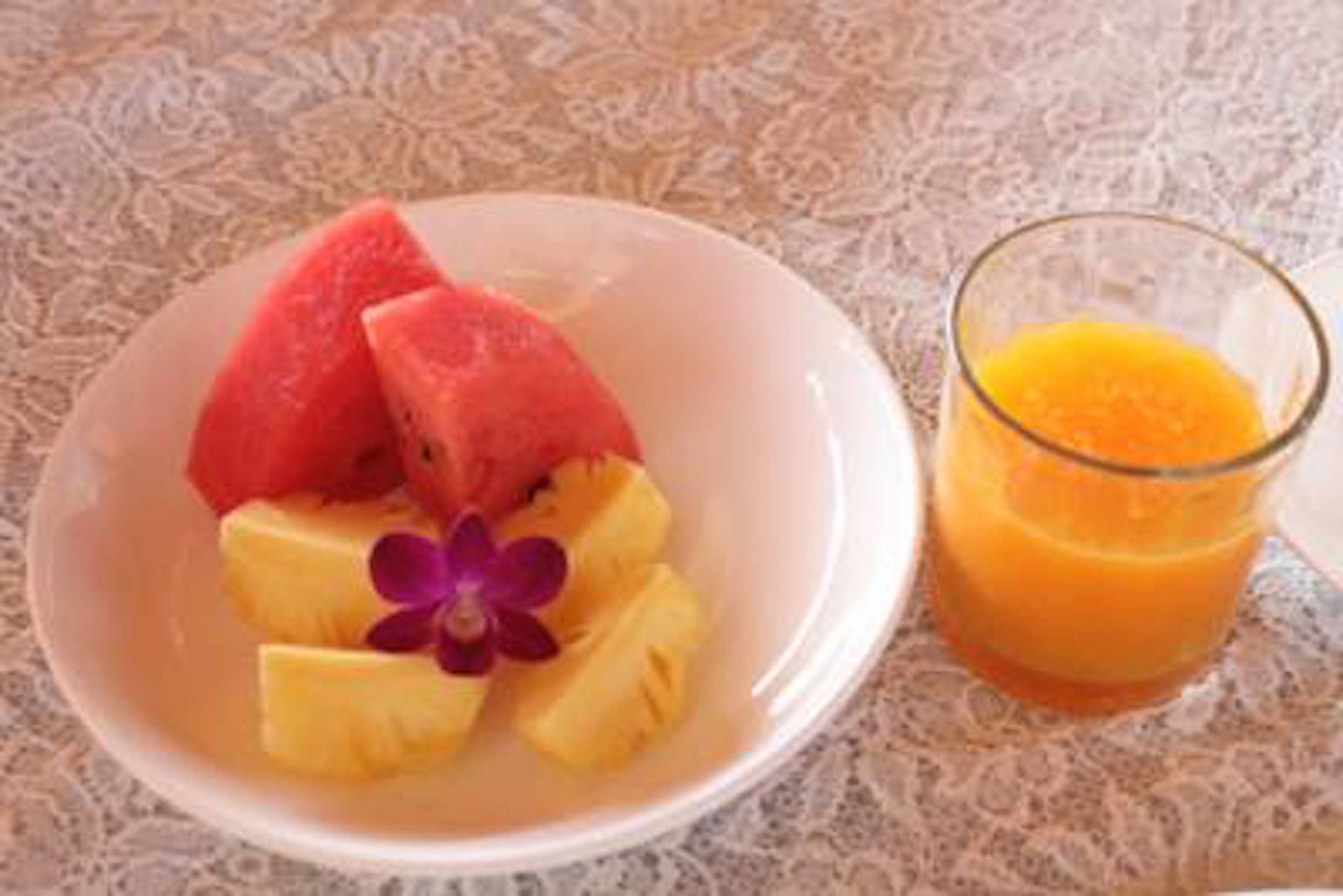 A bowl of fruit with watermelon and pineapple slices, and a glass of orange juice on a textured tablecloth.