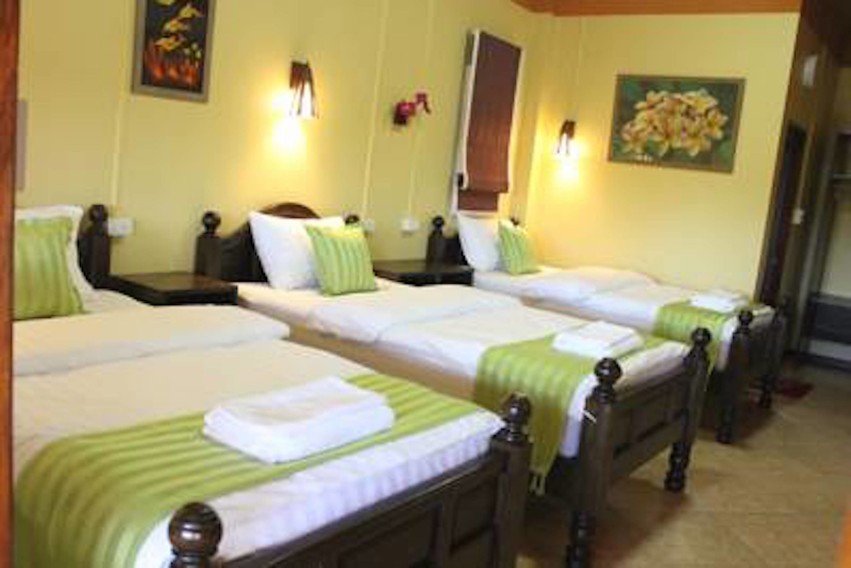 A hotel room with three single beds, each with a wooden headboard and green bedspreads. There are bedside tables with lamps and a large mirror on the wall.