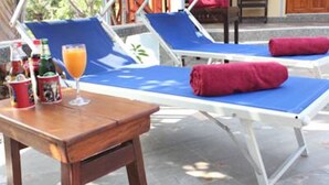 A sunlit outdoor lounge area with blue cushioned chairs, a wooden table with drinks, and a glass of orange juice. - Rimlay Park Resort (Phatthalung)