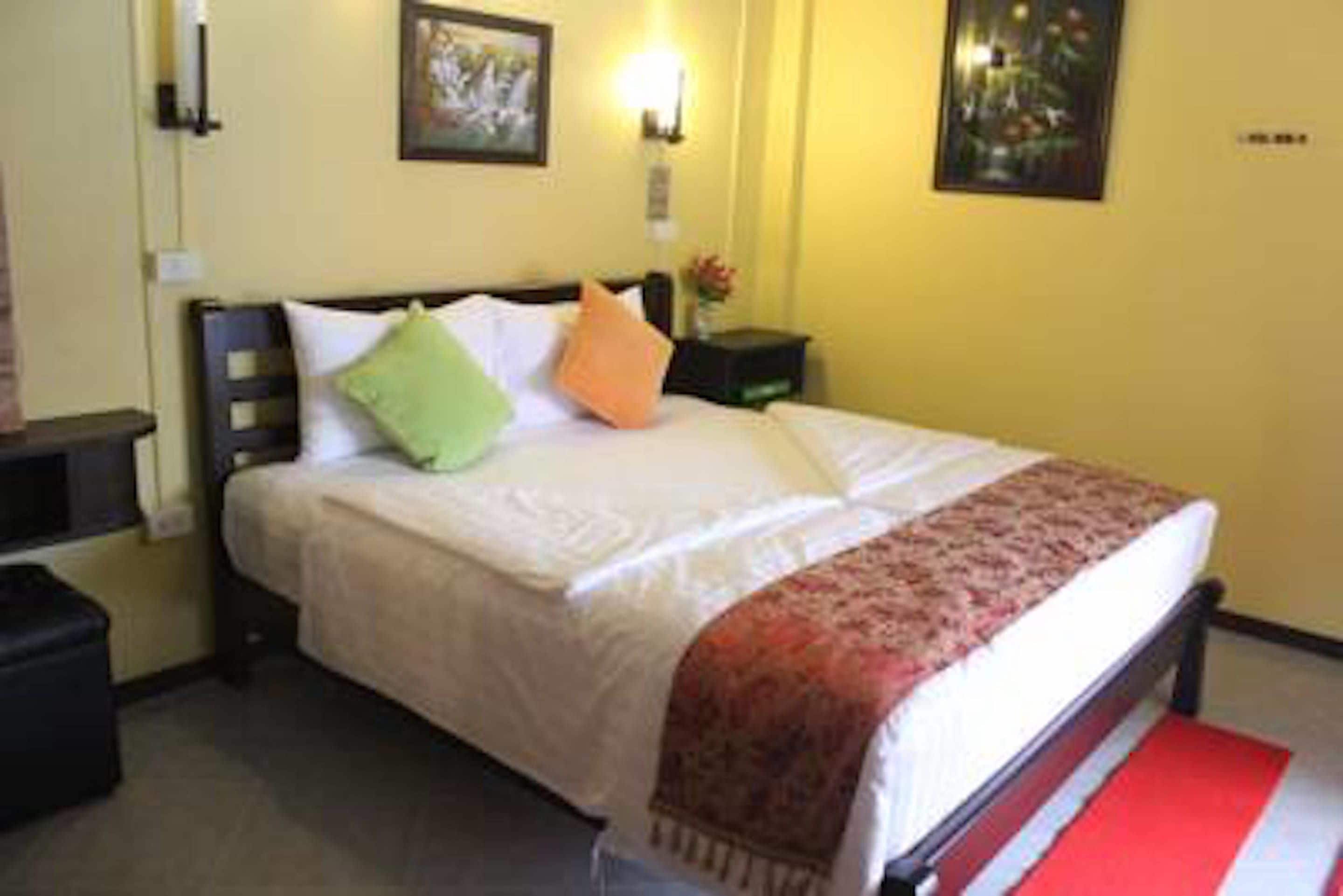 A neatly made bed with a white comforter and a red and white striped runner. There is a green and an orange pillow on the bed. The room has yellow walls and a framed picture on the wall.