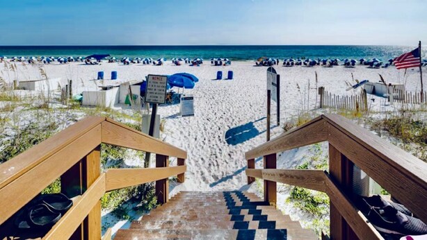Eine Holztreppe führt zu einem Strand mit einem sandigen Pfad, Liegestühlen, Sonnenschirmen und einem Schild.