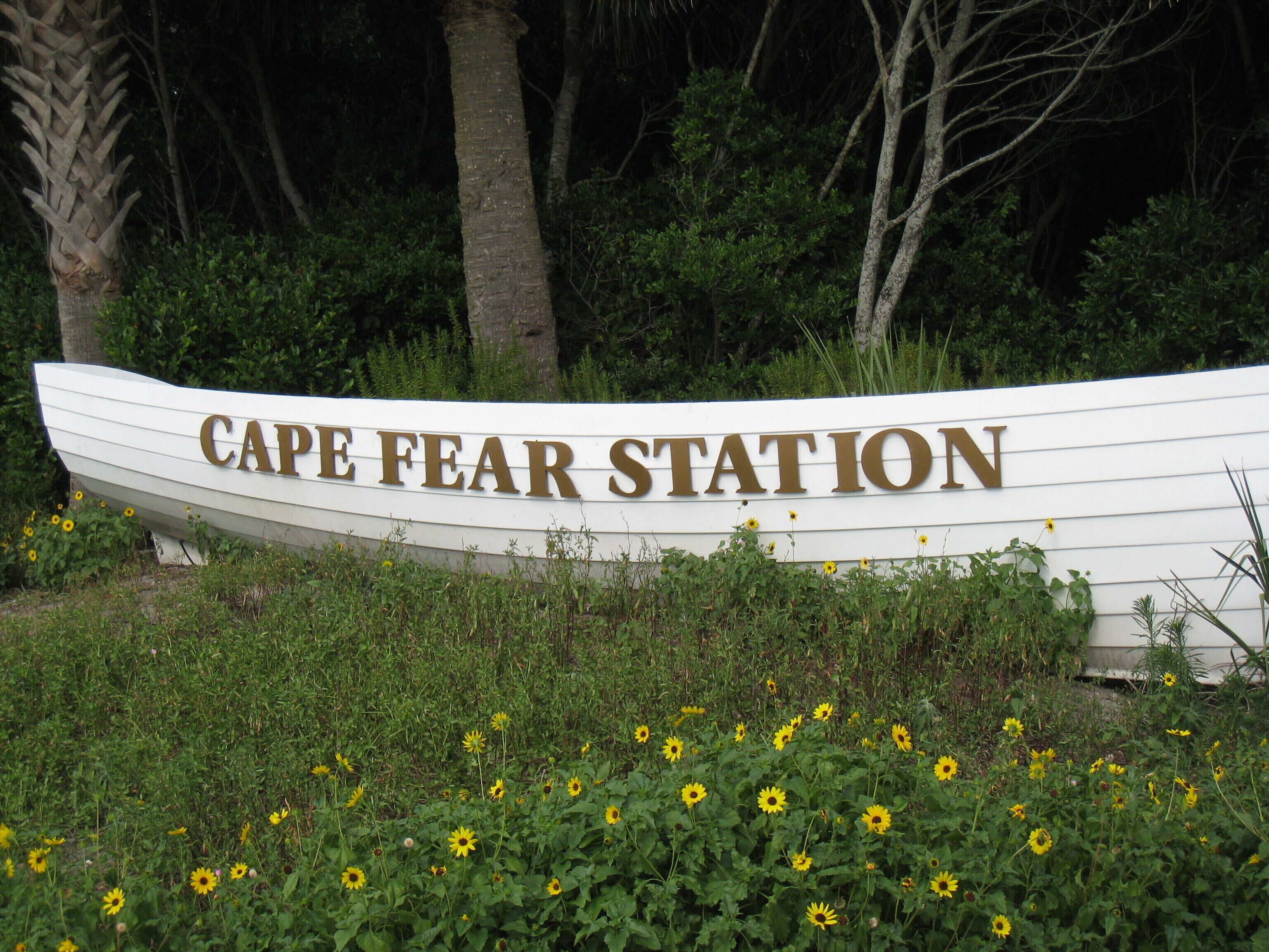A sign with the text "CAPE FEAR STATION" on a white boat-shaped structure surrounded by greenery and sunflowers.