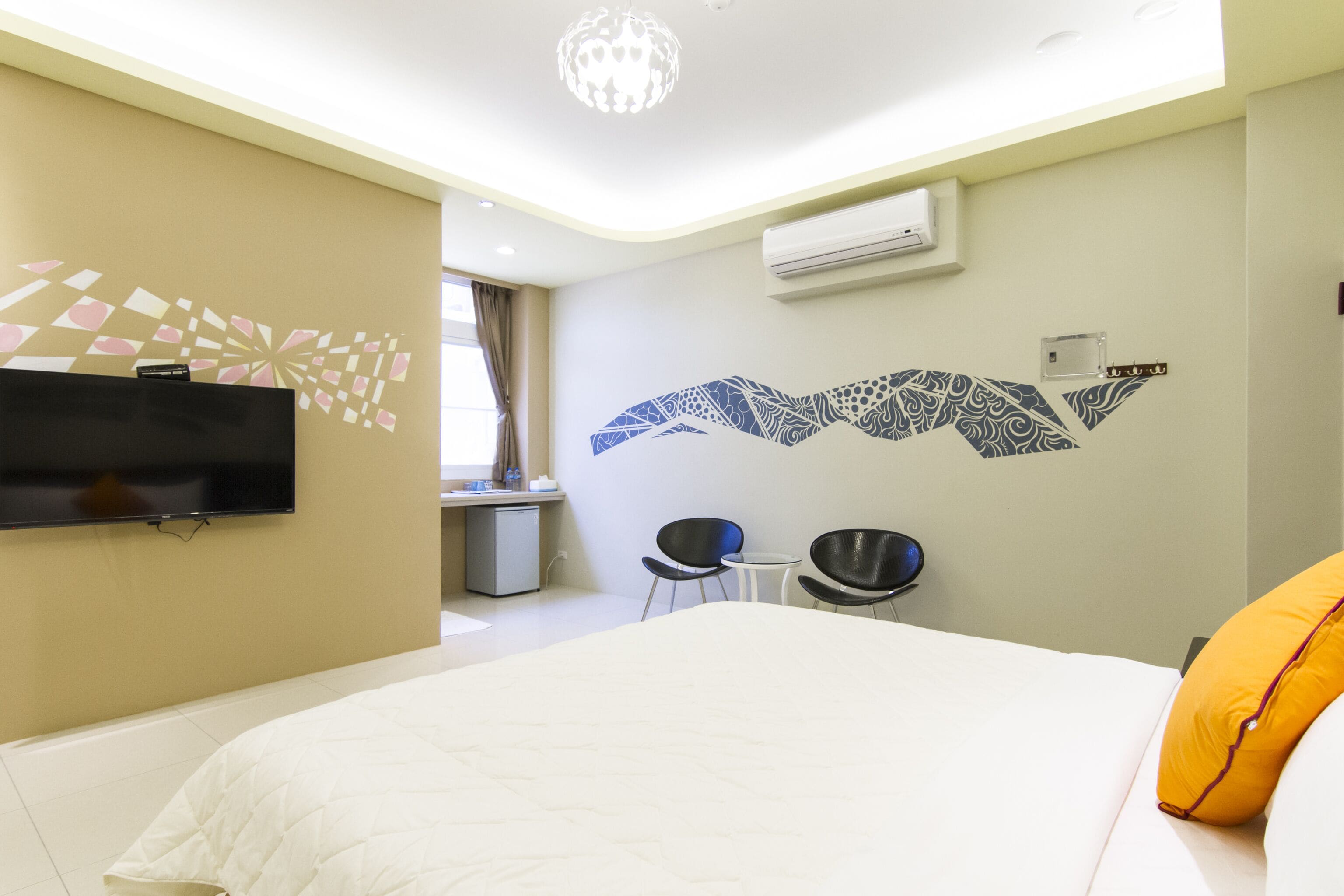 A hotel room with a bed, a flat-screen TV mounted on the wall, two chairs, a small table, and a decorative wall mural.