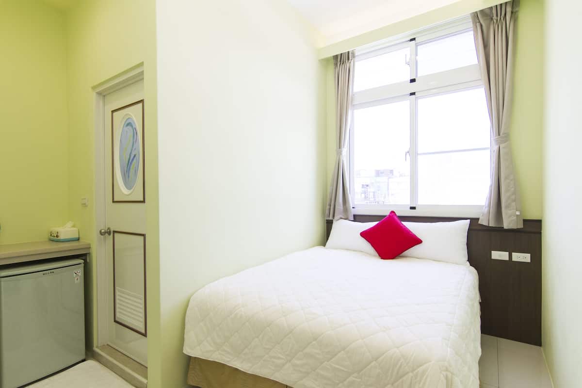 A bedroom with a bed, a window with curtains, a door, and a small kitchenette.