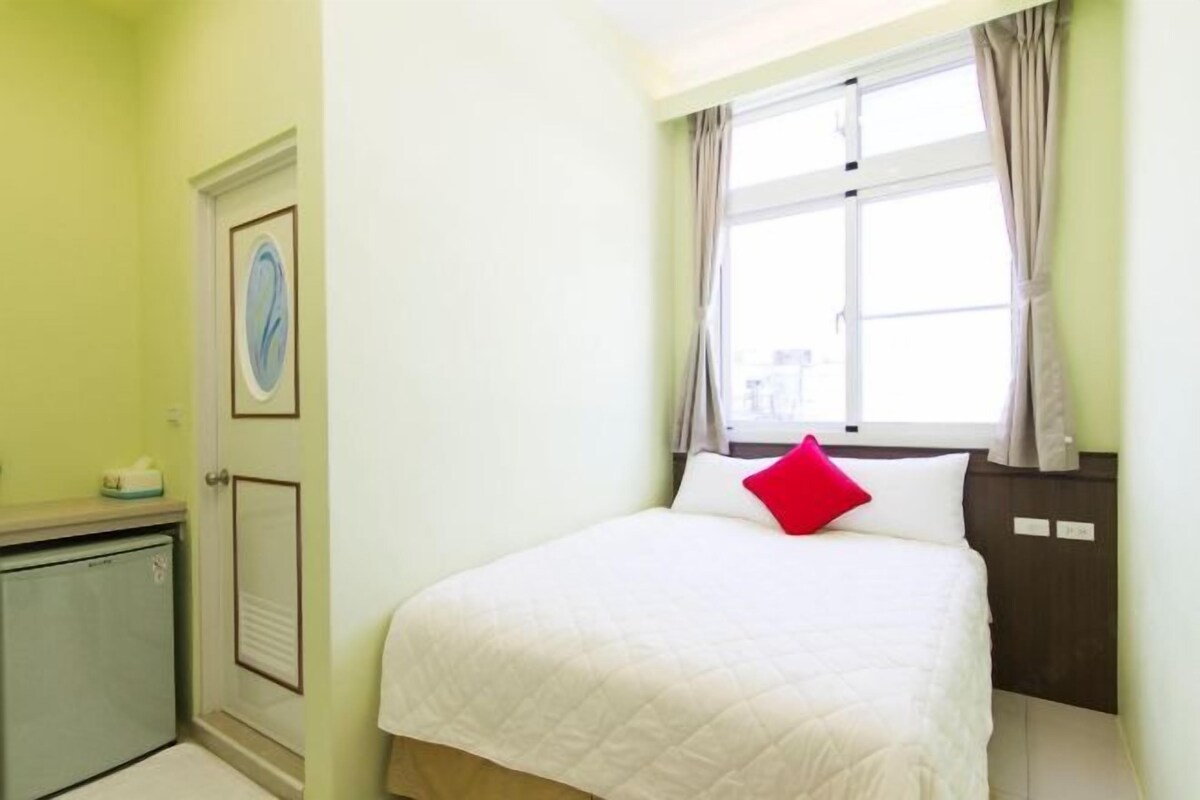 A bedroom with a bed, a window with curtains, a door with a picture, and a small cabinet.