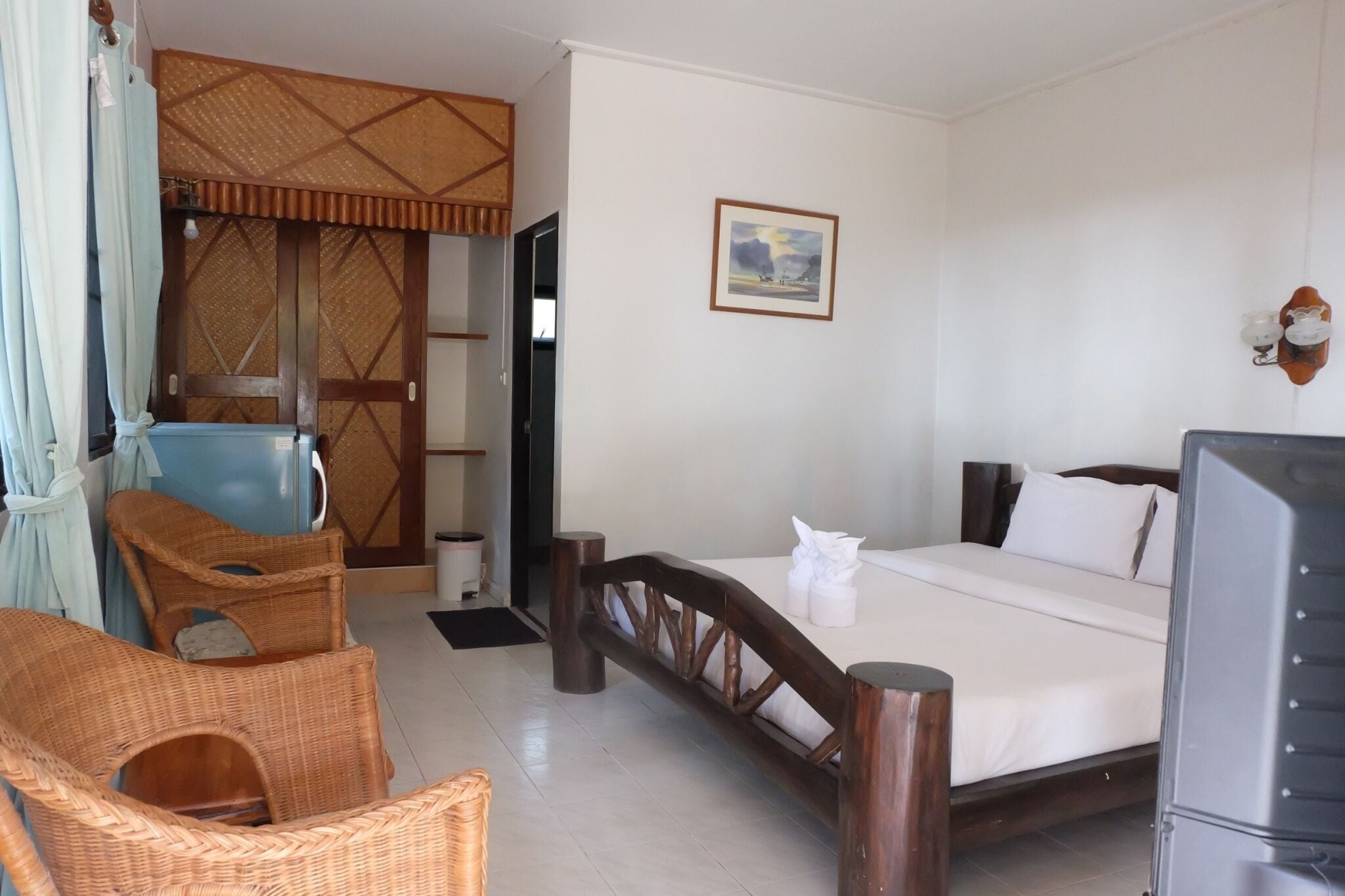 Una habitación de hotel con una cama, televisión, mobiliario de mimbre, una puerta con un diseño decorativo y un cuadro en la pared.