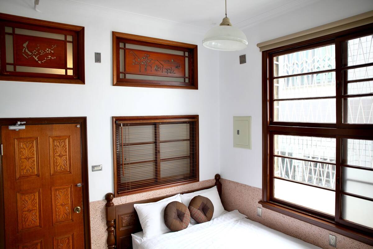 A bedroom with a wooden door, a bed with two pillows, a window with blinds, and a hanging light fixture.