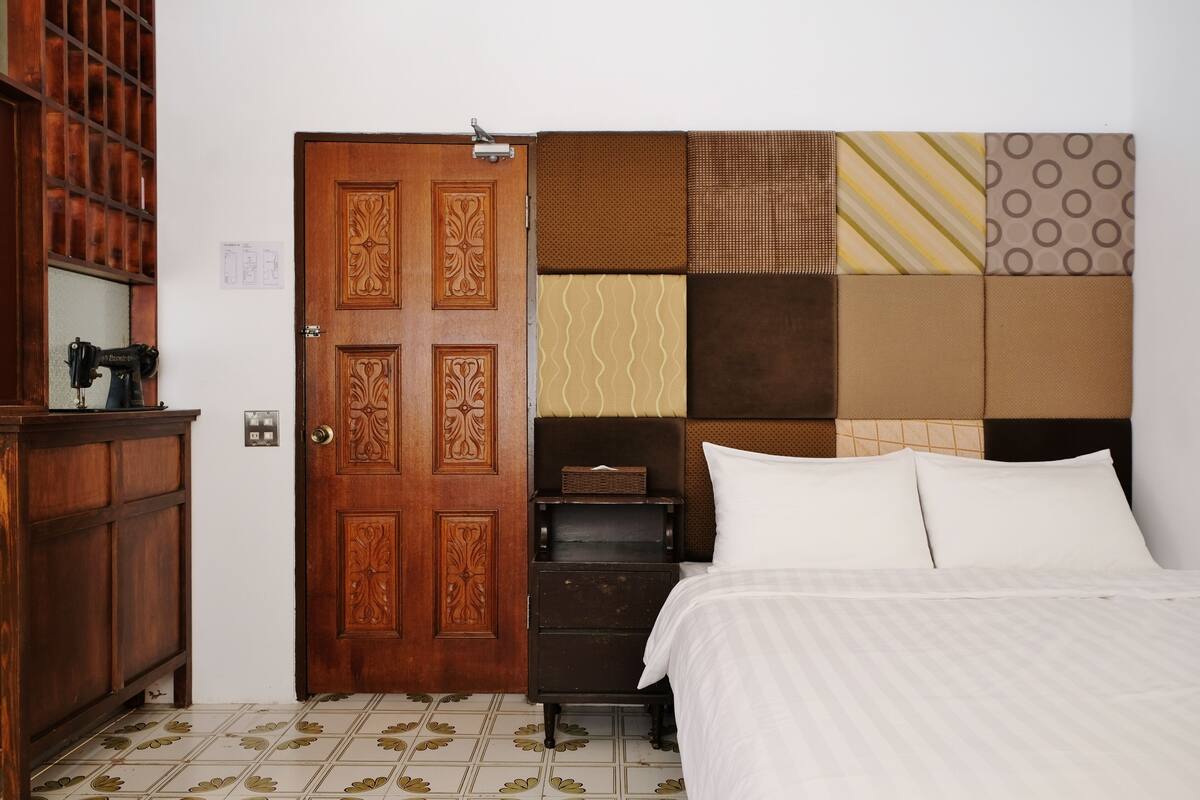 A bedroom with a wooden door, a bed with white bedding, a dresser, and a headboard with patterned tiles.