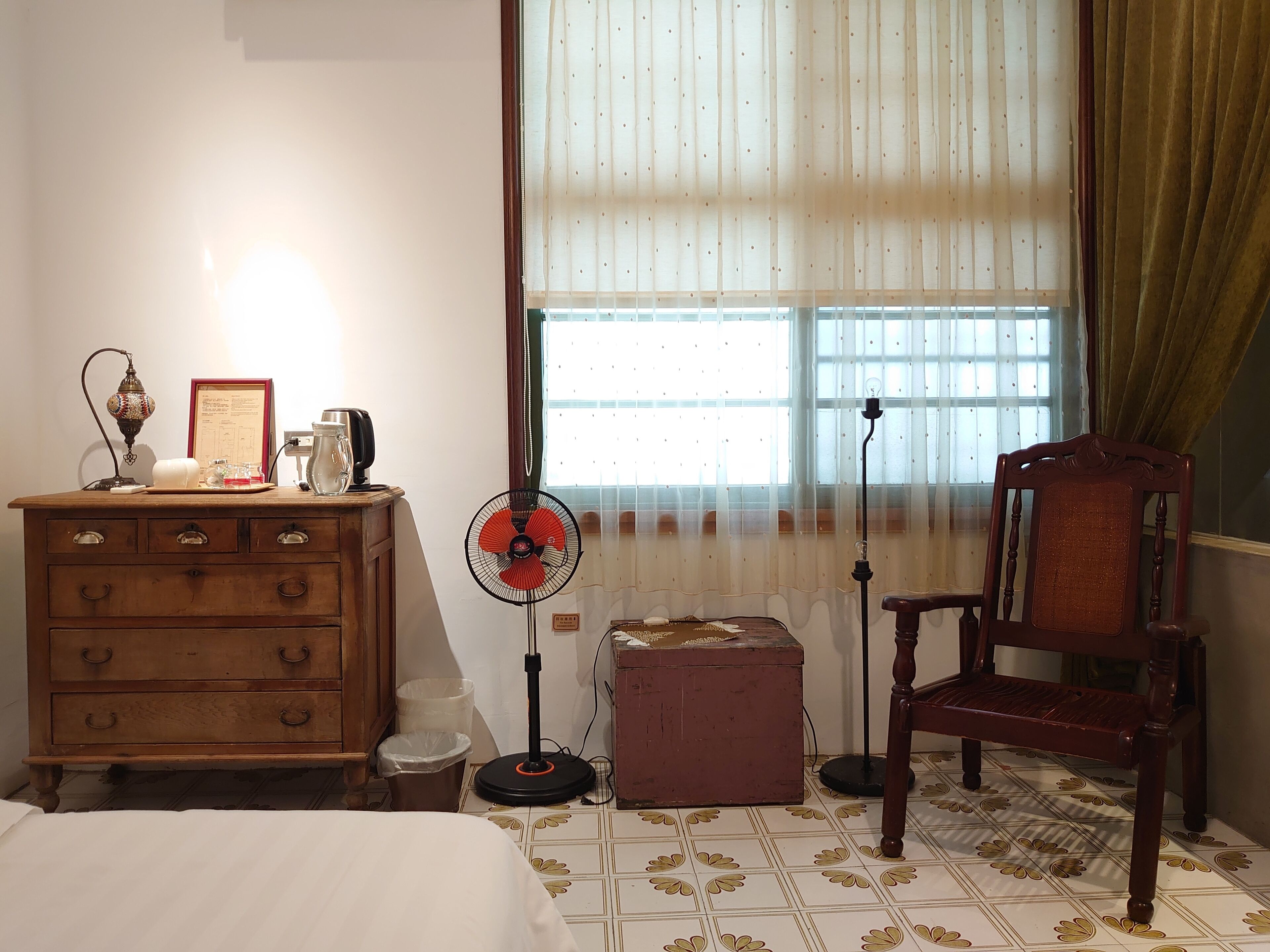 A room with a wooden dresser, a chair, a fan, a lamp, and a window with curtains.