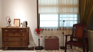 A room with a wooden dresser, a chair, a fan, a lamp, and a window with curtains.