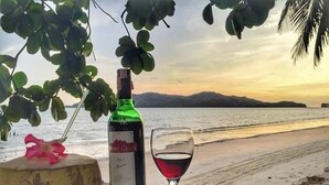 Una botella de vino, una copa de vino tinto y un cuenco de fruta sobre una mesa de madera cerca de la playa.