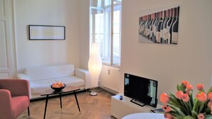 Un salon moderne avec un canapé blanc, une petite table sur laquelle se trouve un bol rempli d’oranges, un téléviseur à écran plat et un vase de fleurs.