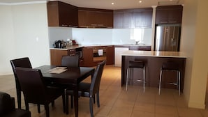 A modern kitchen with dark wooden cabinets, a stainless steel refrigerator, a dining table with four chairs, and a small island with bar stools. - Pier One (Urangan)