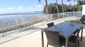 A terrace with a glass railing, wicker chairs, and a table overlooking a coastal view with a pier. - Pier One (Urangan)