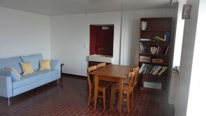 Sala de estar com sofá azul, mesa de jantar de madeira e cadeiras. Há uma estante cheia de livros e uma janela com moldura vermelha.
