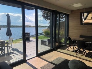 Quarto moderno com grandes portas de vidro deslizantes para um alpendre, área de refeições com cadeiras e vista para um lago.