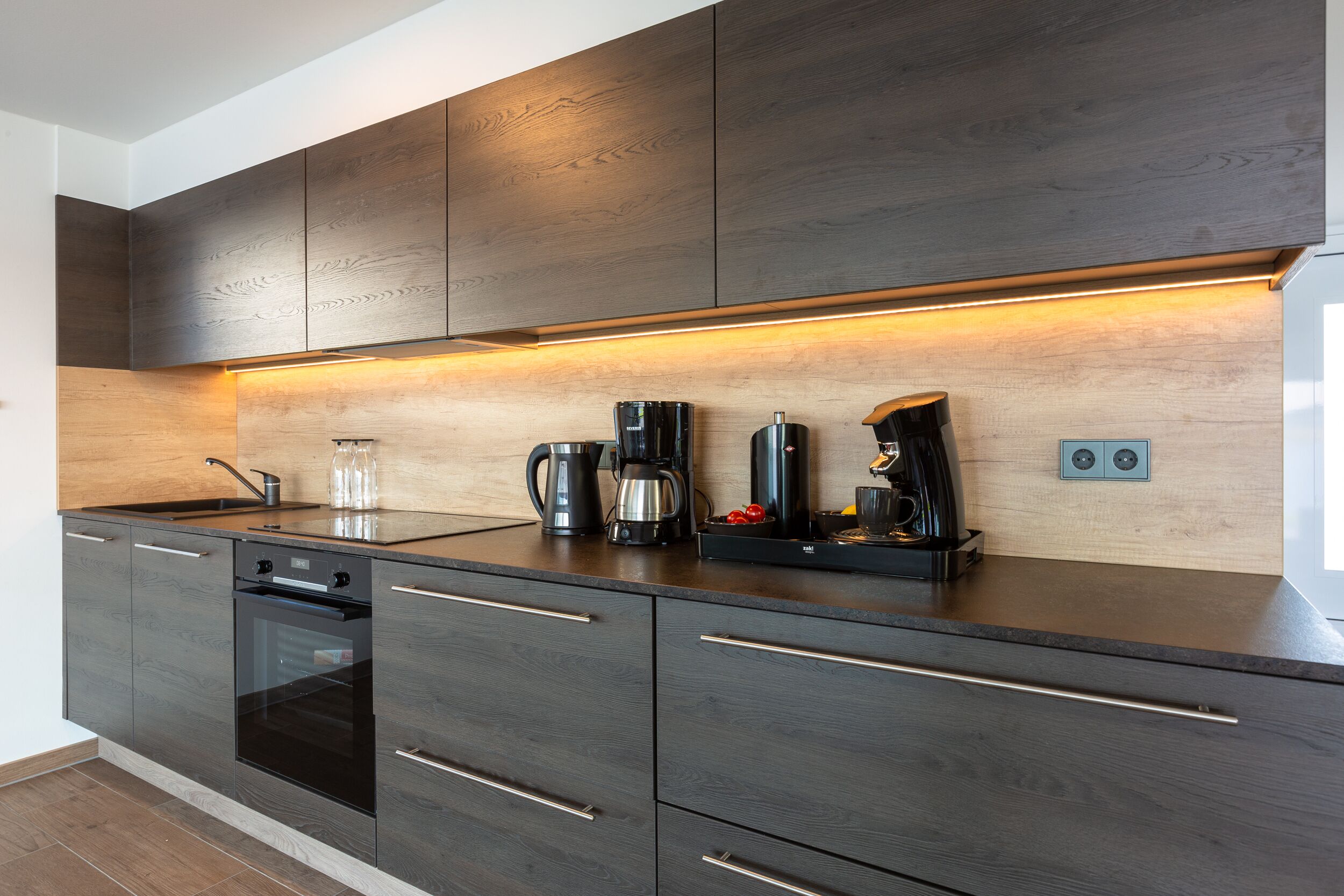 A modern kitchen with dark wood cabinets, a built-in oven, and a coffee station.