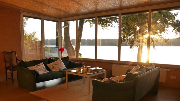 A living room with a green sofa, a coffee table, and a view of a lake through large windows. - Vacation home on Pilsensee with direct lake access, jetty and sauna (Herrsching am Ammersee)