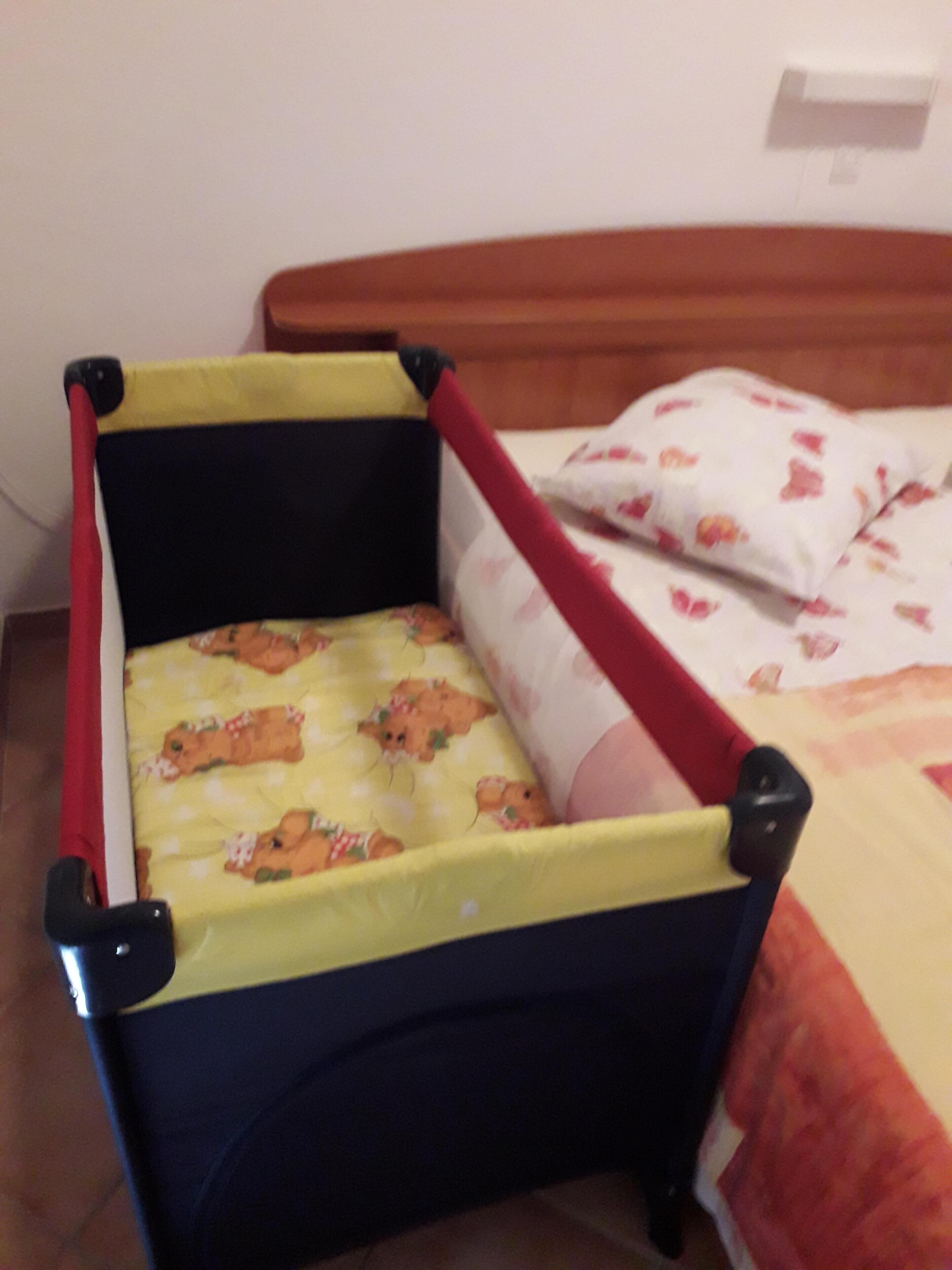 Een kinderbed met een gele matras en een rode rand, geplaatst naast een groter bed met een bloemenprint.