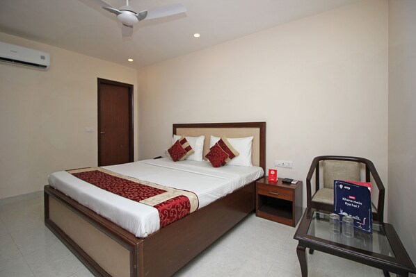 A hotel room with a large bed, a nightstand, a chair, and a table with a sign. - OYO 9028 Hotel Blu Pebbles (New Delhi)