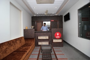 A reception area with a sofa, a desk, a ceiling fan, and a TV mounted on the wall. - OYO 9028 Hotel Blu Pebbles (New Delhi)