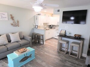 A living room with a grey sofa, a blue coffee table, and a kitchen area with white cabinets and a black TV mounted on the wall.