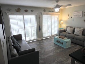 Un salon avec un canapé gris, un divan gris et une table basse bleu clair. Il y a deux fenêtres avec des stores blancs, un ventilateur de plafond et une lampe fixée au mur.