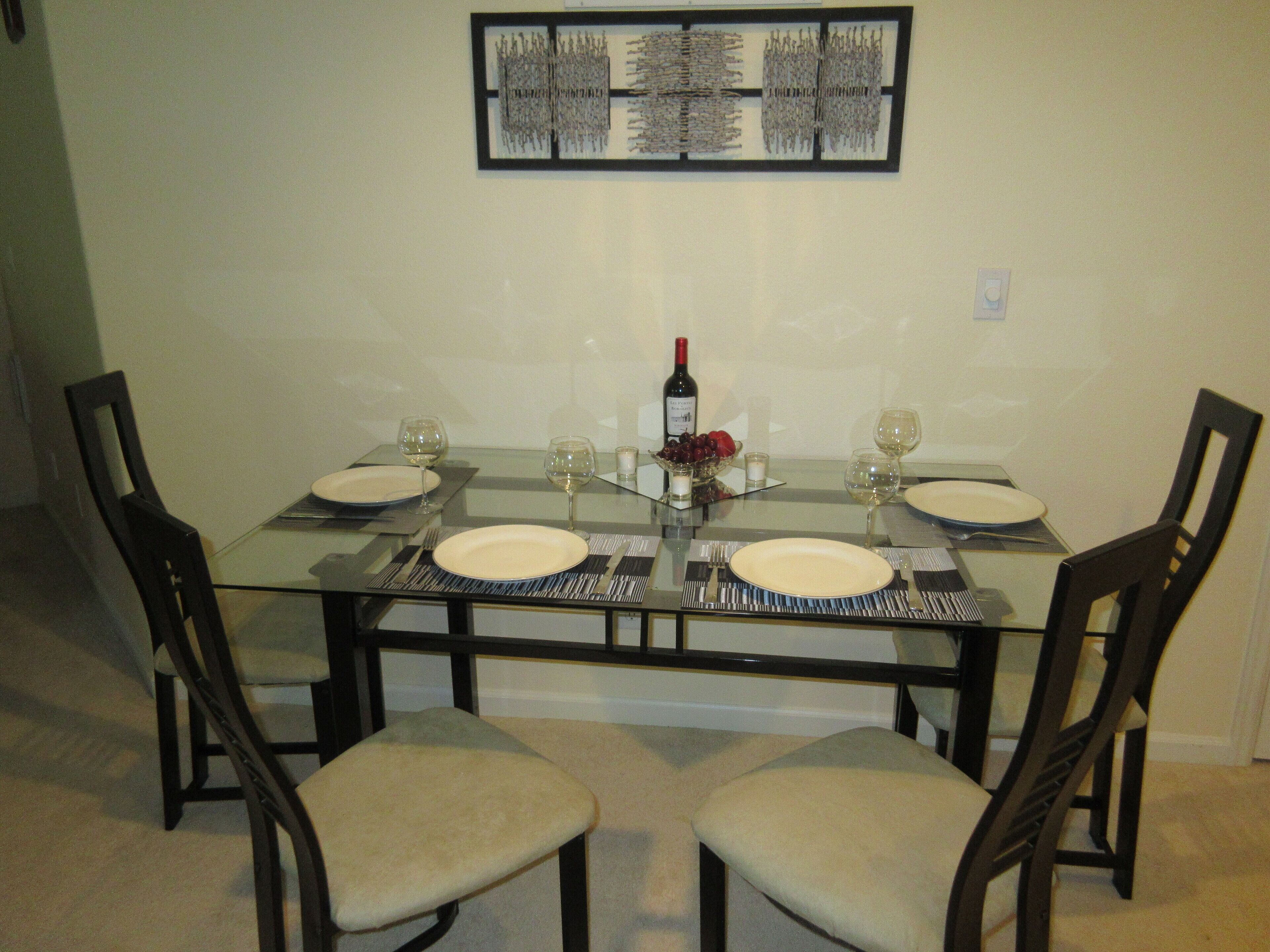 Mesa de comedor lista para cuatro personas, con sillas, una botella de vino y copas.