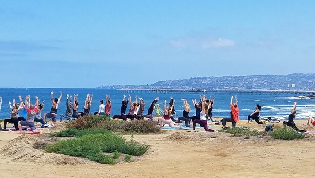 A group of people performing yoga on a beach with a pier in the background.