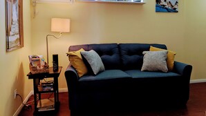 A living room with a dark blue sofa, a small table with books, a lamp, and decorative items on a shelf above the sofa.