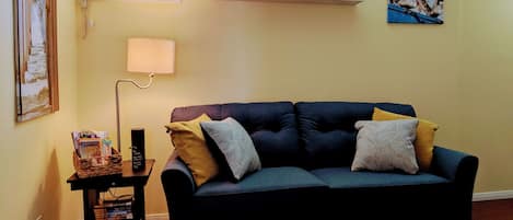 Ein Wohnzimmer mit einem dunkelblauen Sofa, einem kleinen Tisch mit Büchern, einer Lampe und Dekoration auf einem Regal über dem Sofa.