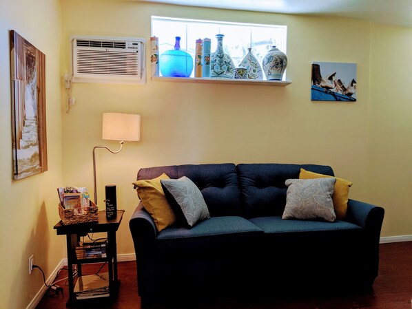 A living room with a dark blue sofa, a small table with books, a lamp, and decorative items on a shelf above the sofa.