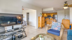 A modern living room with a flat-screen TV, a coffee table, and a wicker chair. - Maui Banyan - Ocean View G Condo (Kihei)