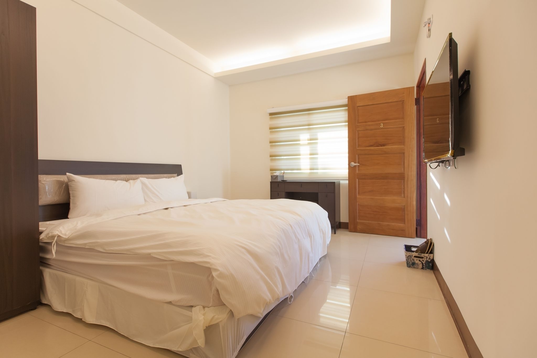 Een slaapkamer met een bed, een houten deur, een klein bureau en een aan de muur bevestigde televisie.