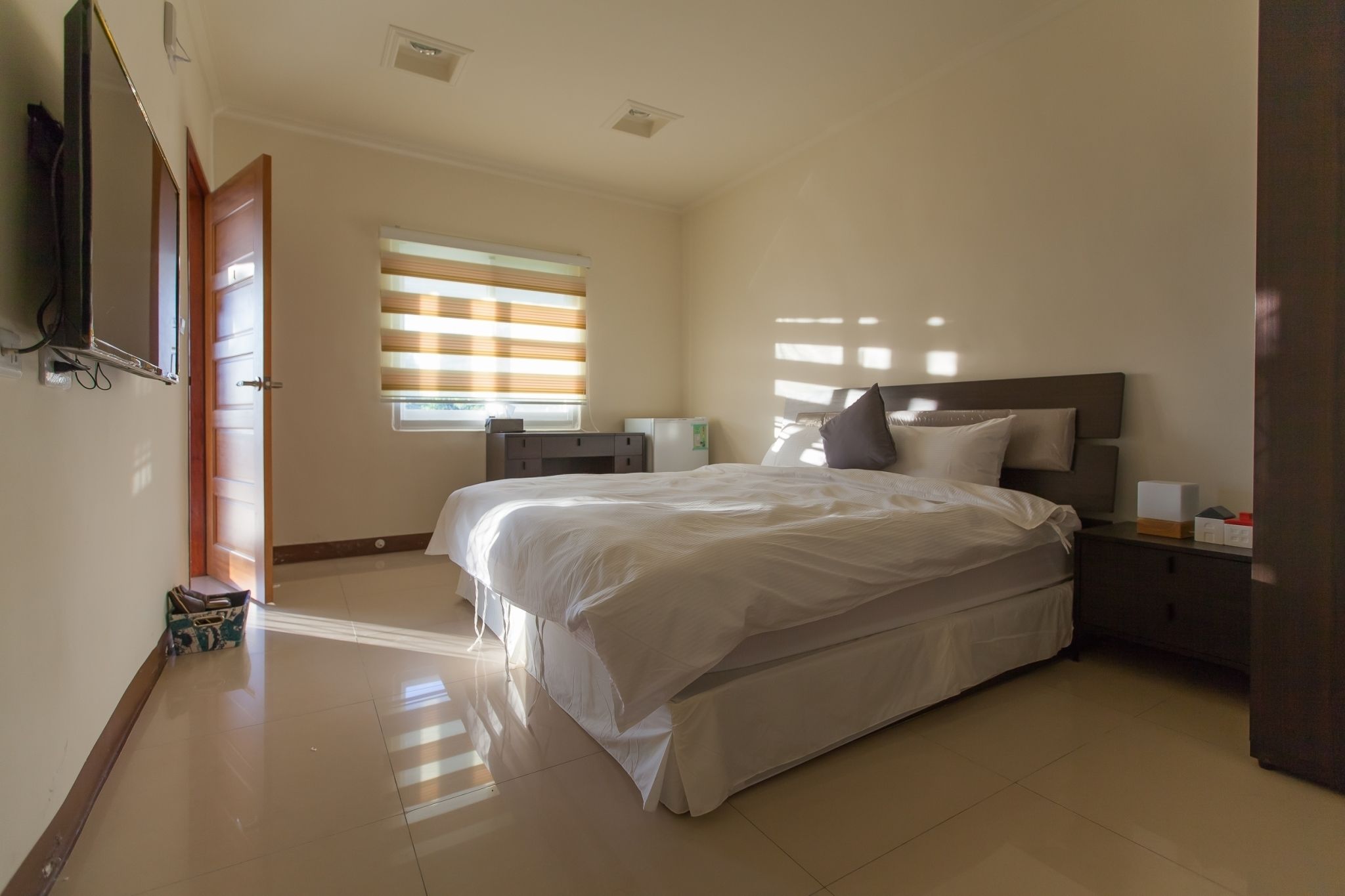 A hotel room with a large bed, a bedside table, a TV mounted on the wall, and a window with blinds.