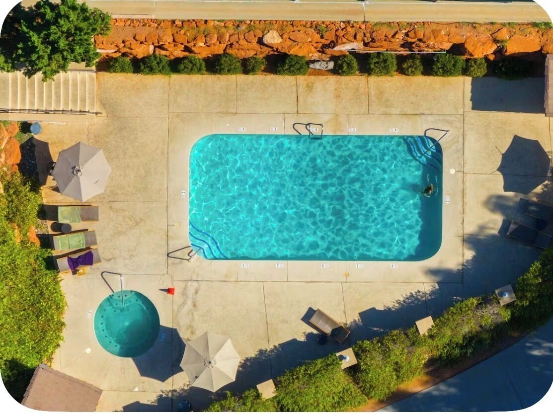 A rectangular swimming pool with a clear blue surface, surrounded by a paved deck with lounge chairs and umbrellas.