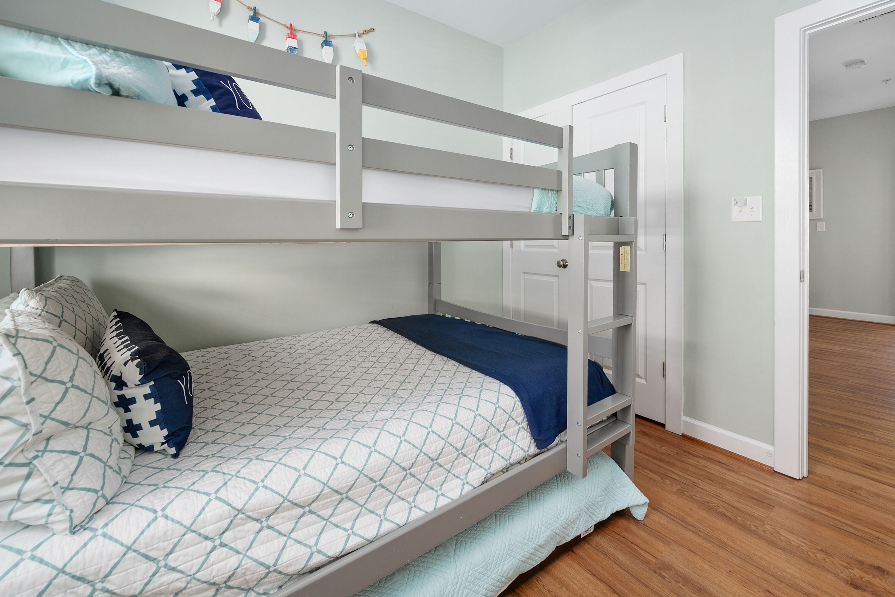 Una cama litera con colcha acolchada y almohadas, piso de madera y una puerta que da a otra habitación.