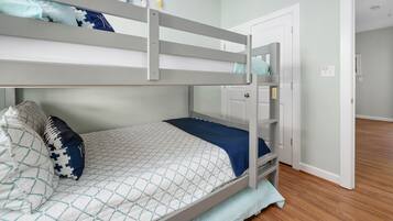 Una cama litera con colcha acolchada y almohadas, piso de madera y una puerta que da a otra habitación.