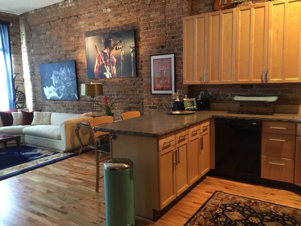 A modern kitchen with wooden cabinets, a central island, and a brick wall. There is a dining area with a sofa and a coffee table.