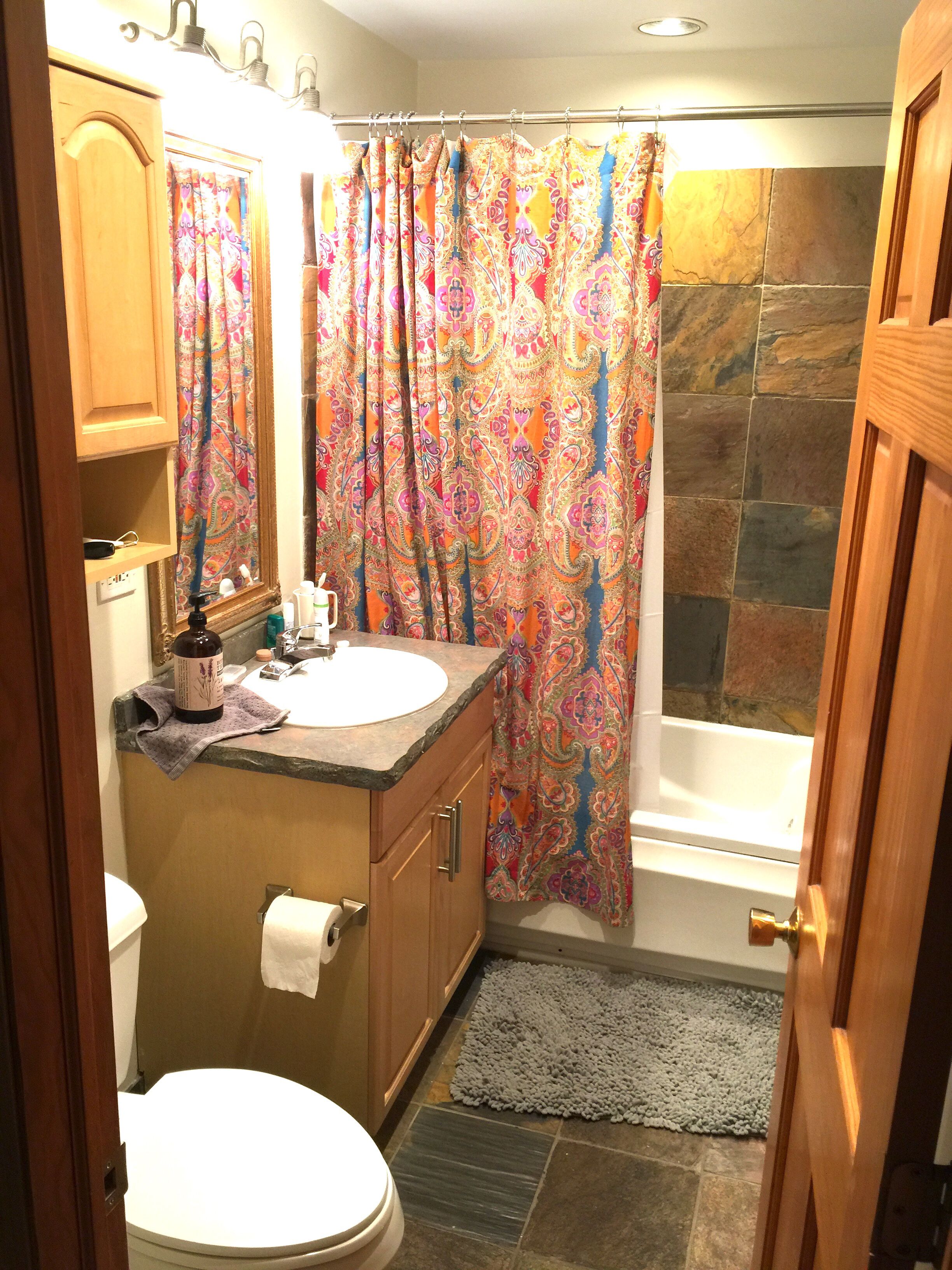 A bathroom with a wooden vanity, a toilet, a bathtub, and a shower curtain with a paisley pattern.