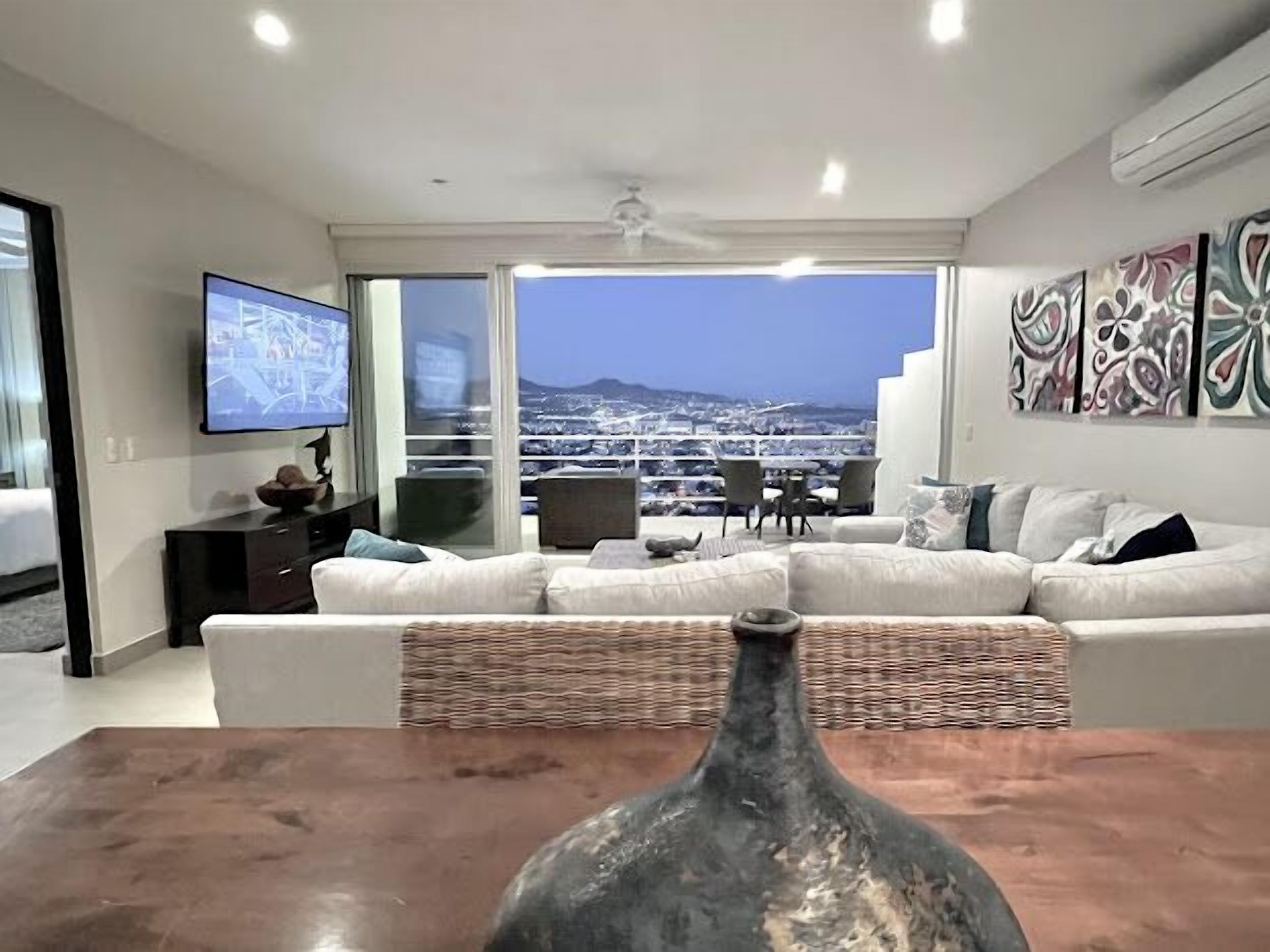 A modern living room with a large TV, a dining table with a vase, and a balcony with a city view.