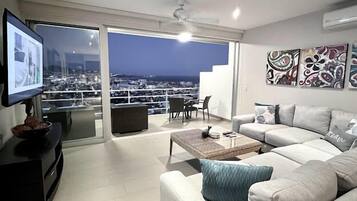 A modern living room with a sofa, coffee table, and a television. There is a balcony with a city view.