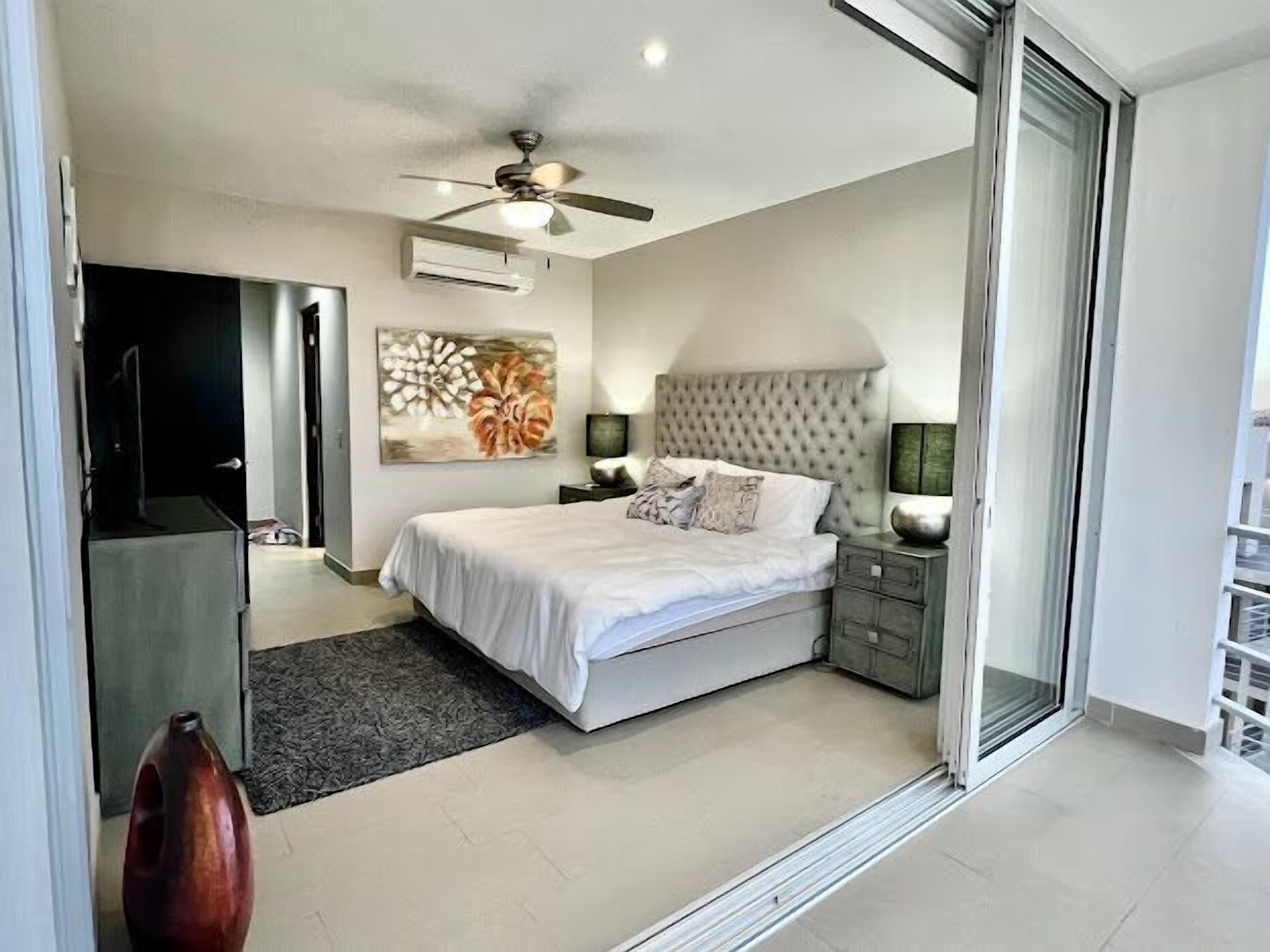 A modern bedroom with a large bed, a ceiling fan, an air conditioning unit, a decorative painting, and a nightstand with a lamp.