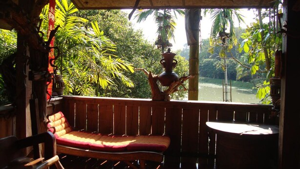 A wooden bench with a red cushion, a small table, and a decorative pot on a balcony with a view of a river and surrounding greenery. - Baan Suan Jantra Home Stay (Chiang Rai)